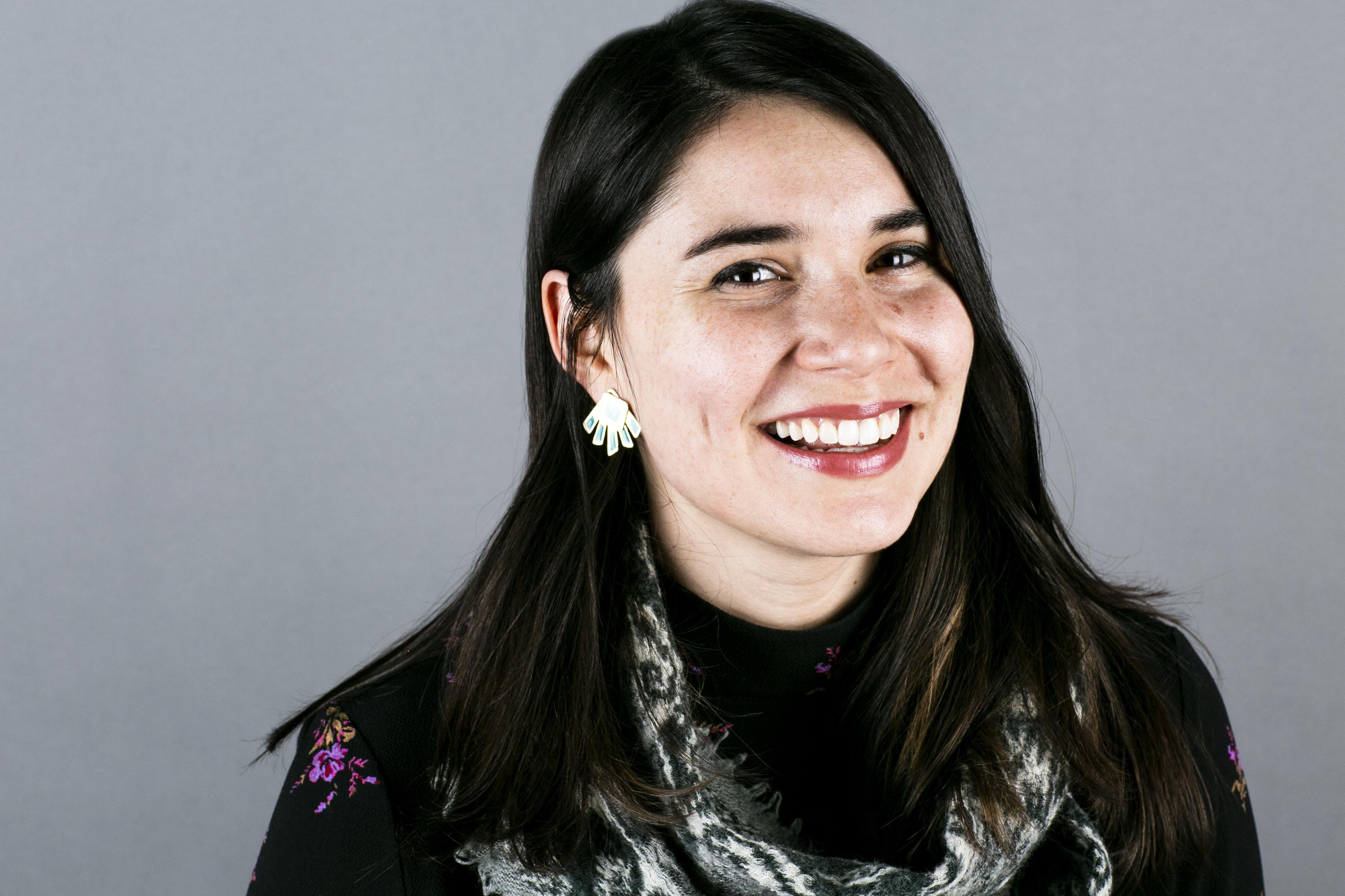 Woman, Gabriella Garcia-Pardo, smiling in a studio portrait, with dark hair, wearing patterned earrings and a scarf against a gray background.