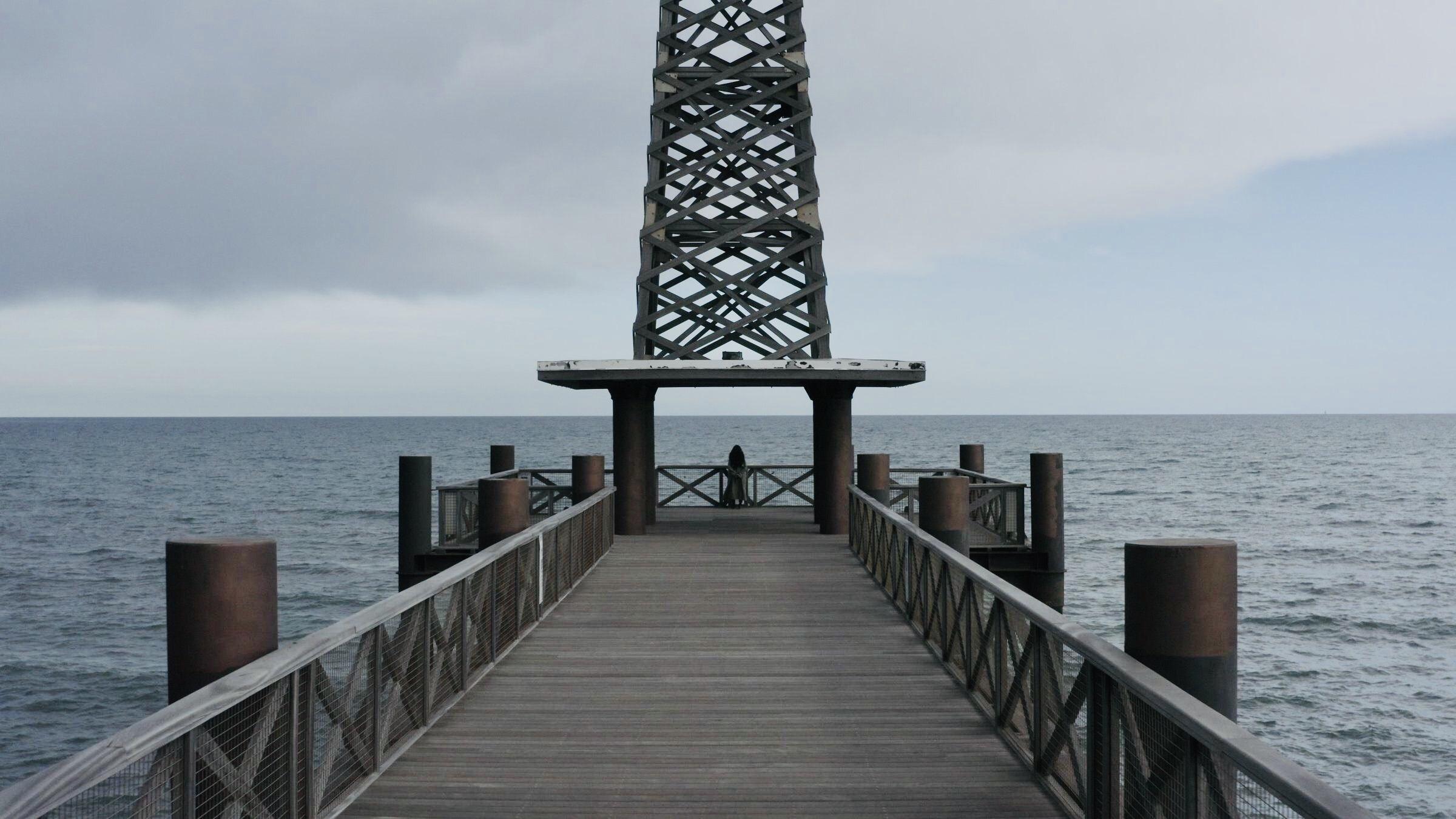 Still from Eskape. A person stands at the end of a wooden pier, facing the ocean. The pier leads to a structure resembling a small observation deck with a lattice tower.