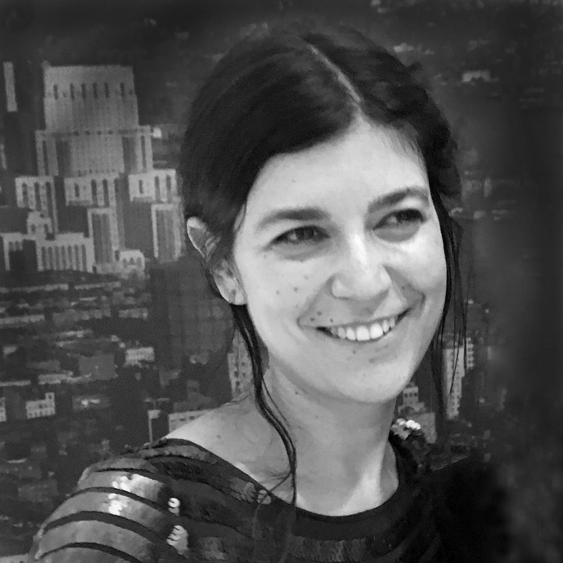 Rachel Elizabeth Seed is behind the lens of a camera on a tripod. The camera aims at a landscape and the sun is setting. Portrait in black and white.