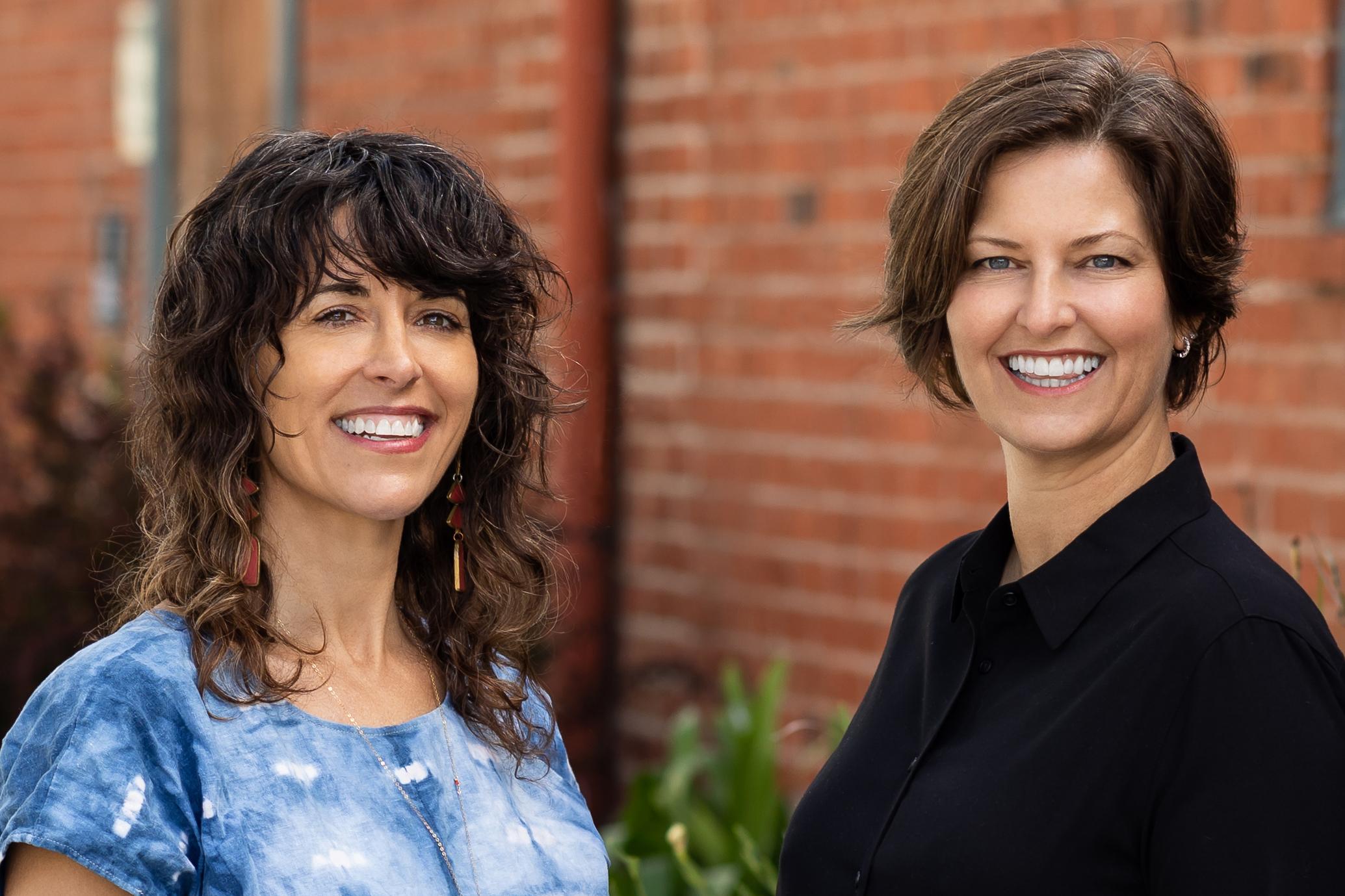 Two women are pictured together standing outside in front of a brick wall.
Left: Rivkah, a white woman with shoulder length curly hair stands in a blue and white blouse smiling.
Right: Jen, a white woman with short brown hair, stands in a black top smiling.