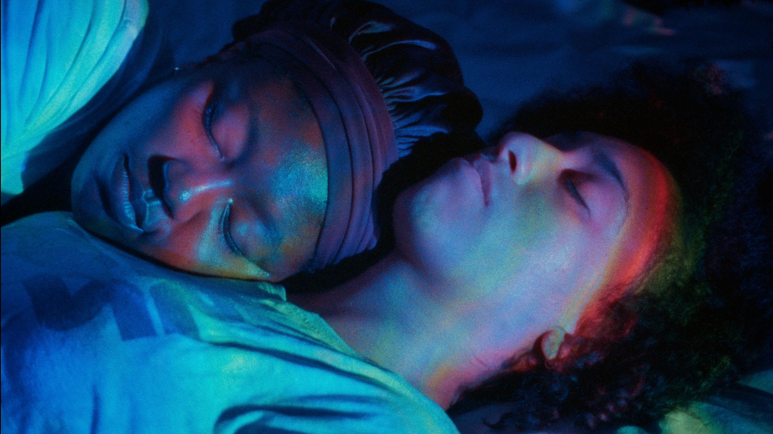 Two women sleep cuddled up. Blue and purple lighting falls on their faces.