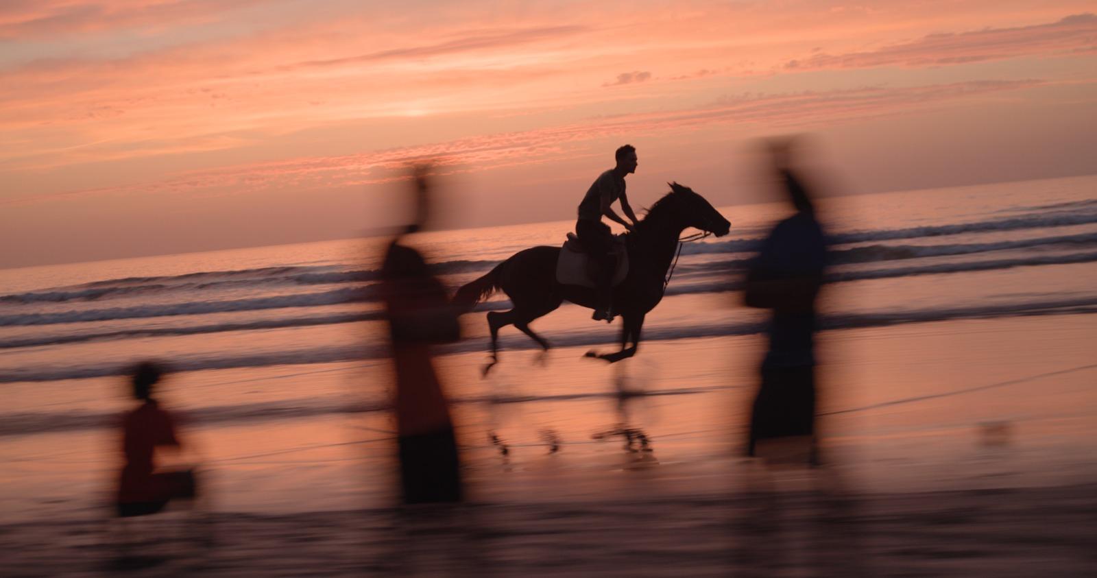 A man gallops a horse on a beach at sunset as blurry figures watch in the foreground.