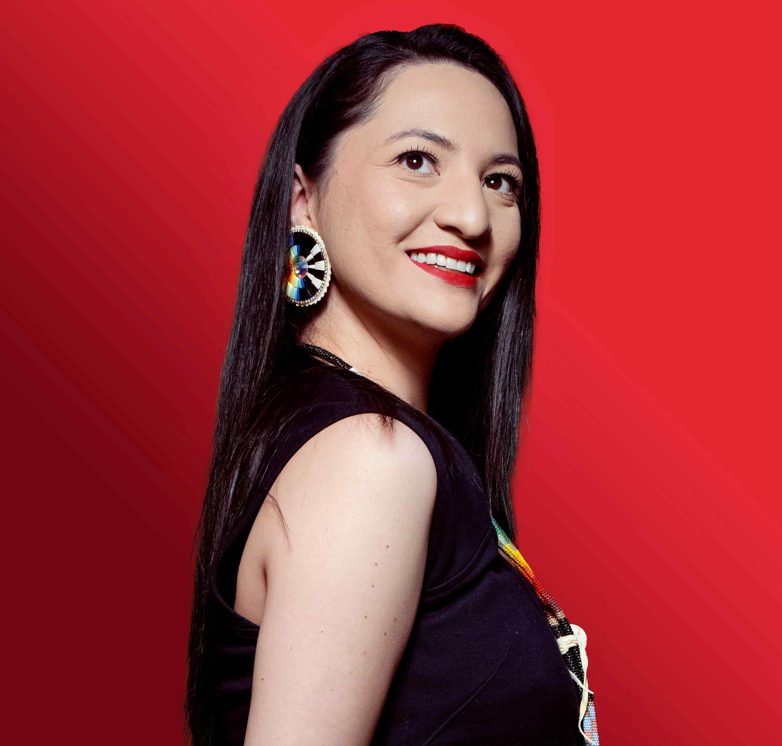Tsanavi Spoonhunter stands in front of a red background with a smile looking above the camera. She is wearing a black shirt with a matching set of quill earrings and a beaded lanyard.