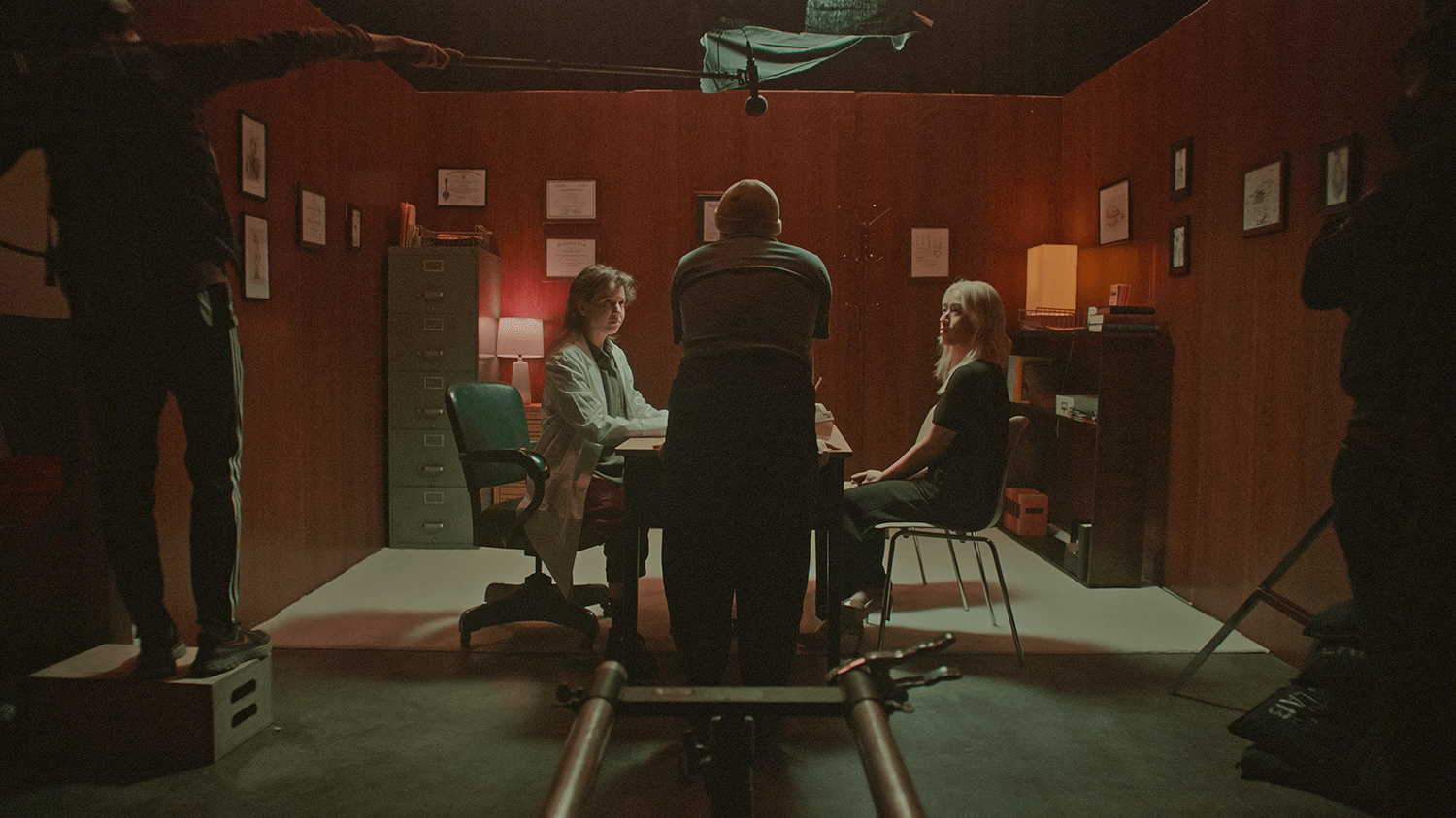 Still from The Script. In a set, a person with their back to the camera, talks to two other persons that are sitting in chairs in front of a table. To the right and left, there are camera crew people. In a dimly-lit set that resembles a doctor's office, two actors, dressed as a doctor and patient, look intently at the director speaking to them. The Script is a short film in the QUEER FUTURES series.