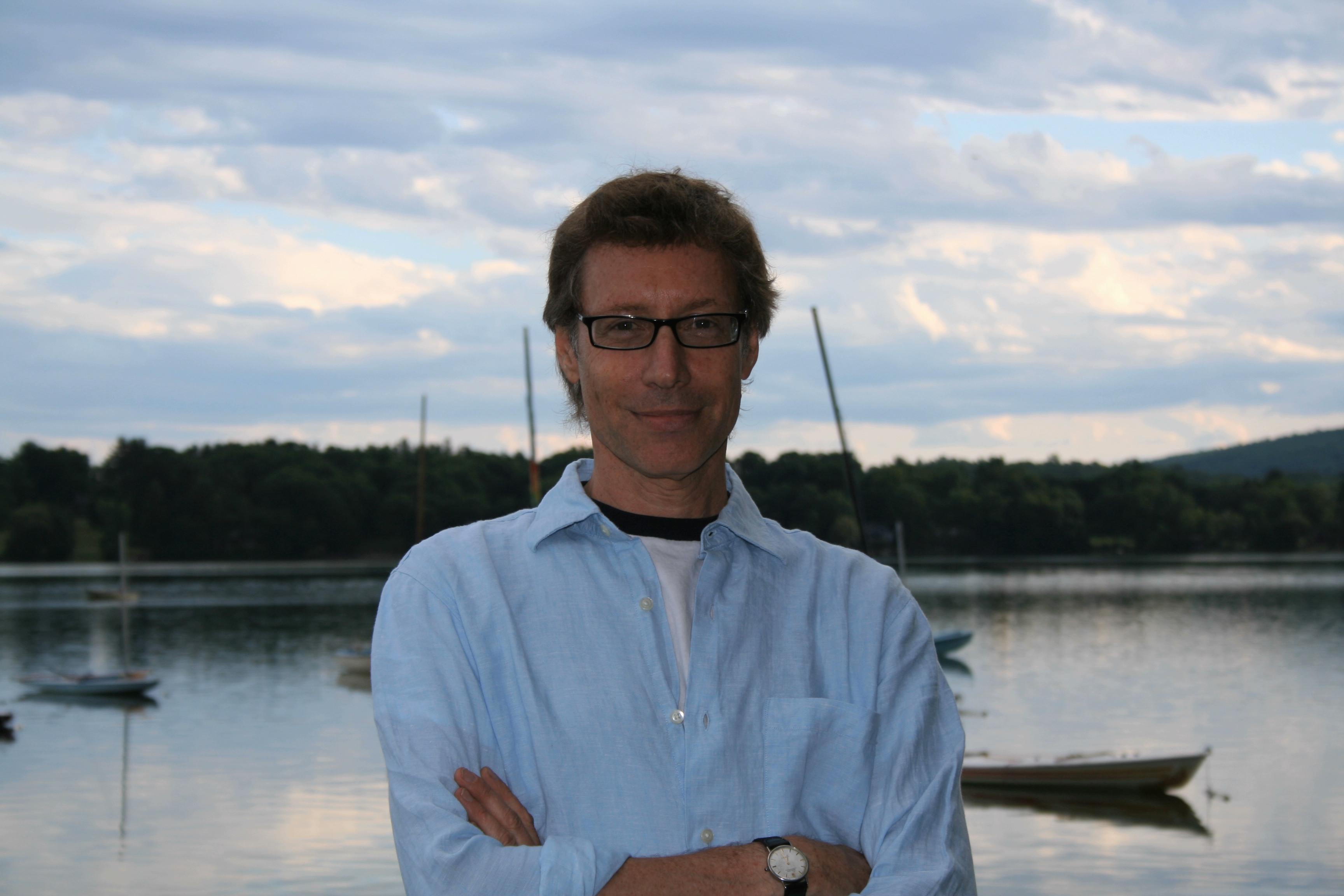 Man with glasses smiles at the camera in a blue shirt. He has short brown hair.
