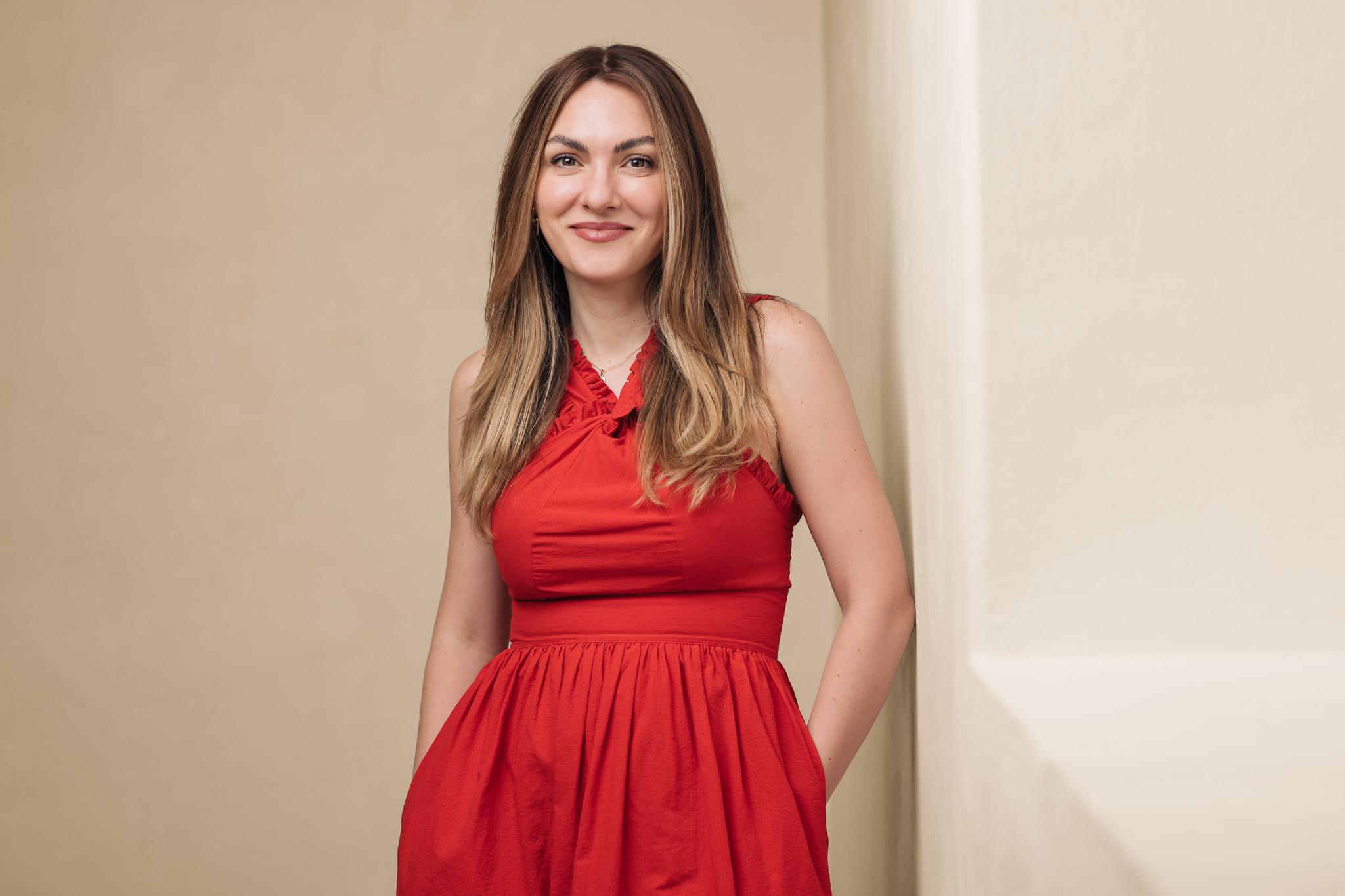 Portrait of a woman wearing a red, formal dress, standing in front of a beige background and smiling into the camera.