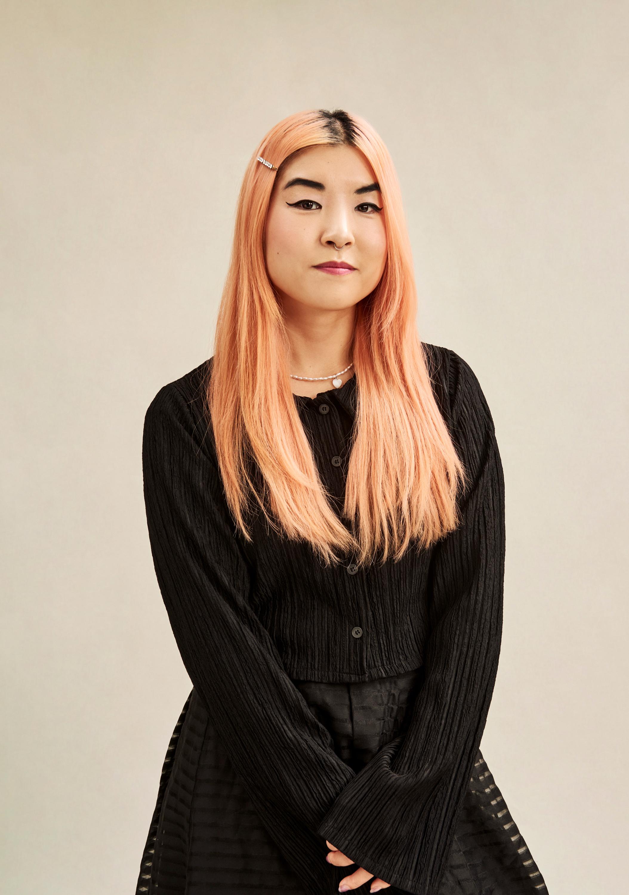 Director So Yun Um stands on a beige background in an all-Black outfit.