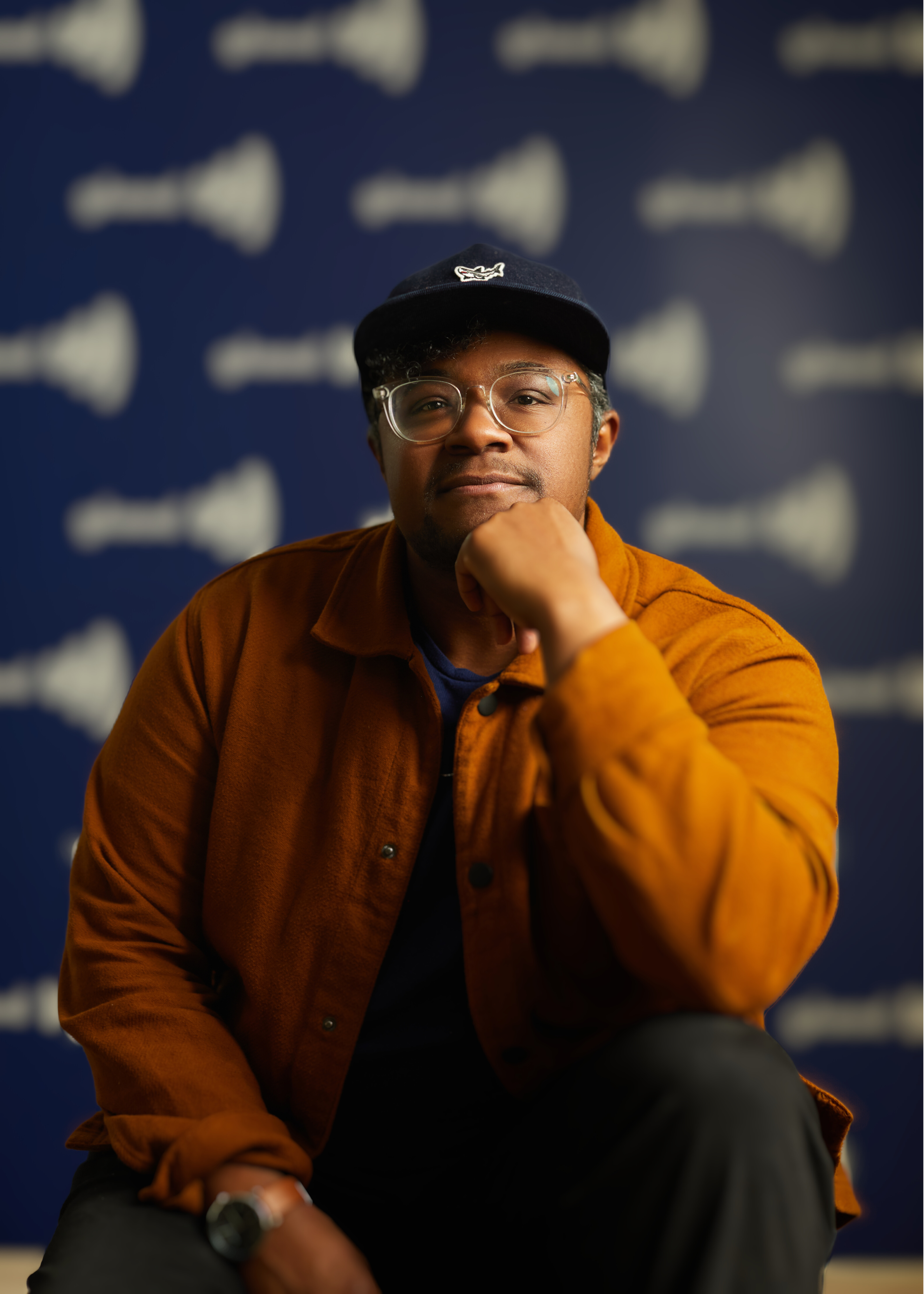 A person wearing glasses, a black cap, and an orange jacket sits with one hand resting on their chin against a blue background with white abstract patterns.