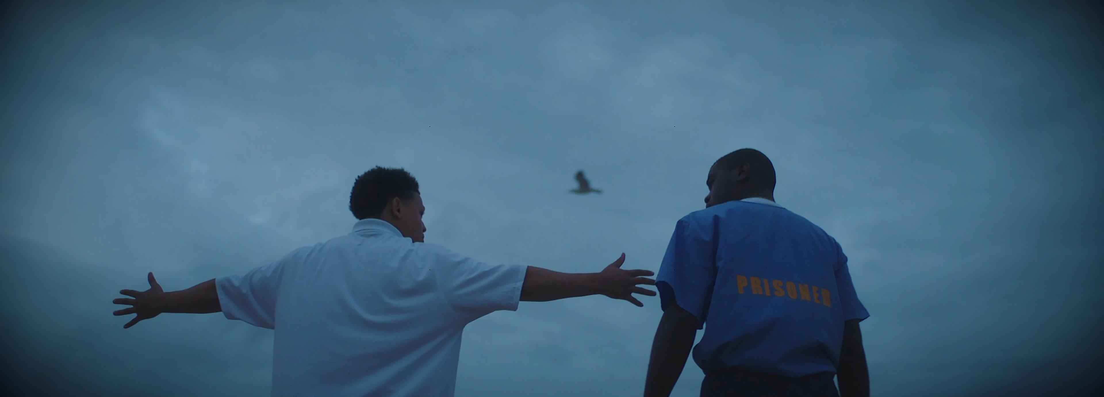 At dusk, two men stand next to each other while a bird flies overhead. One man stretches his arms out wide as he speaks, the other wears a prison uniform.