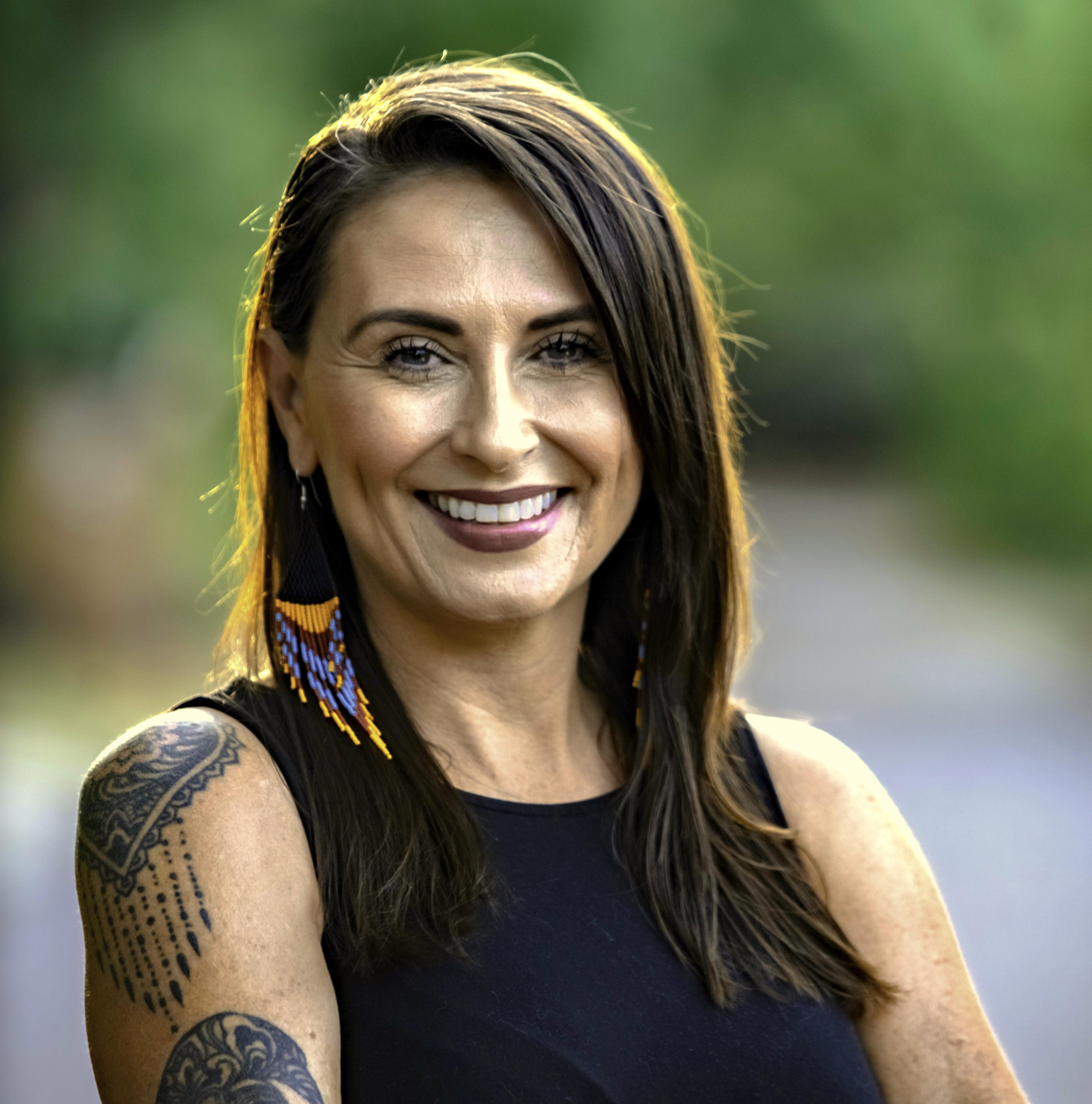 Portrait of Julianna Brannum smiling outdoors, with long brown hair, wearing a black sleeveless top and colorful beaded earrings, with a tattoo visible on her upper arm. Soft green foliage fills the blurred background.