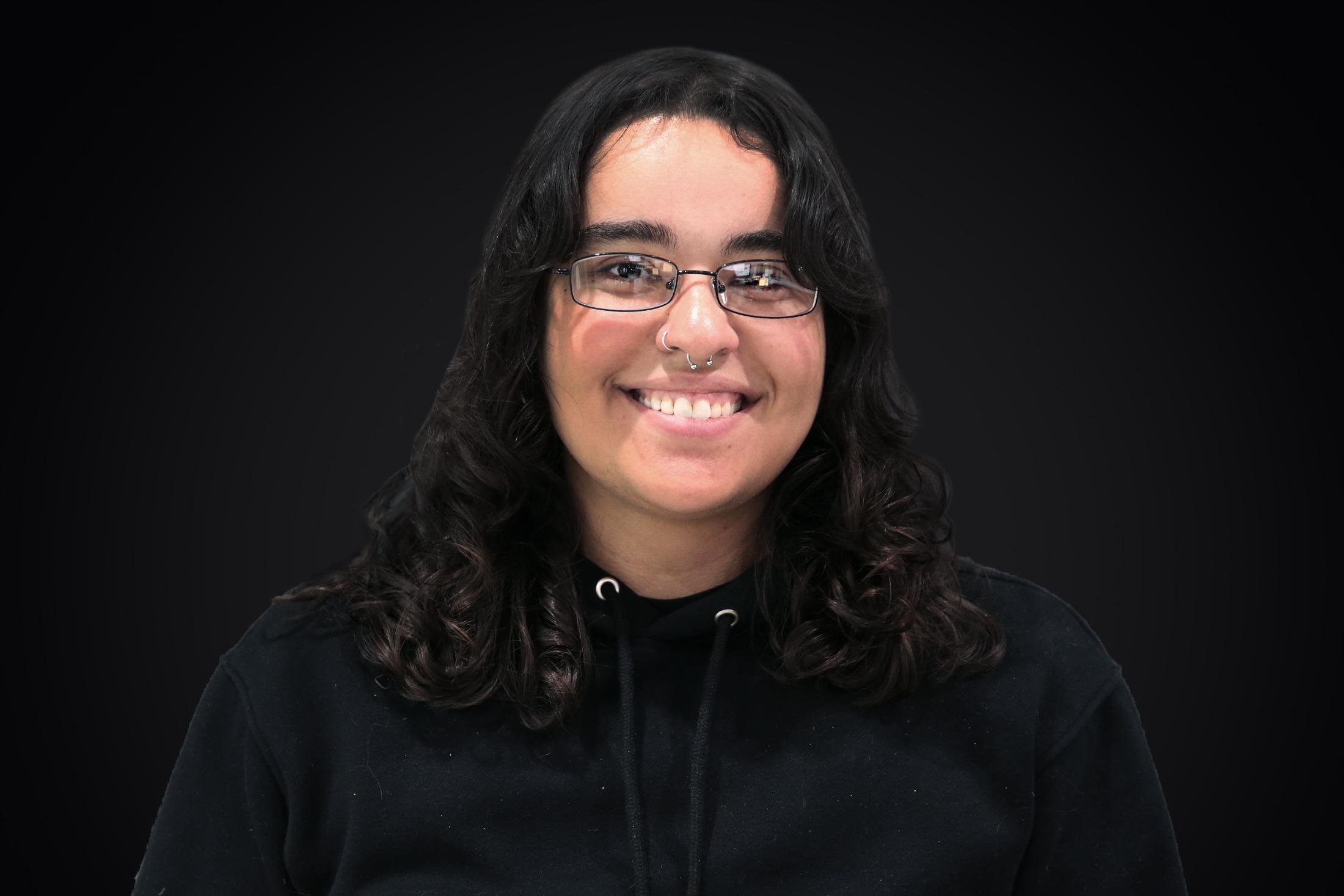 A woman with glasses and curly hair smiles at the camera in front of a dark backdrop.