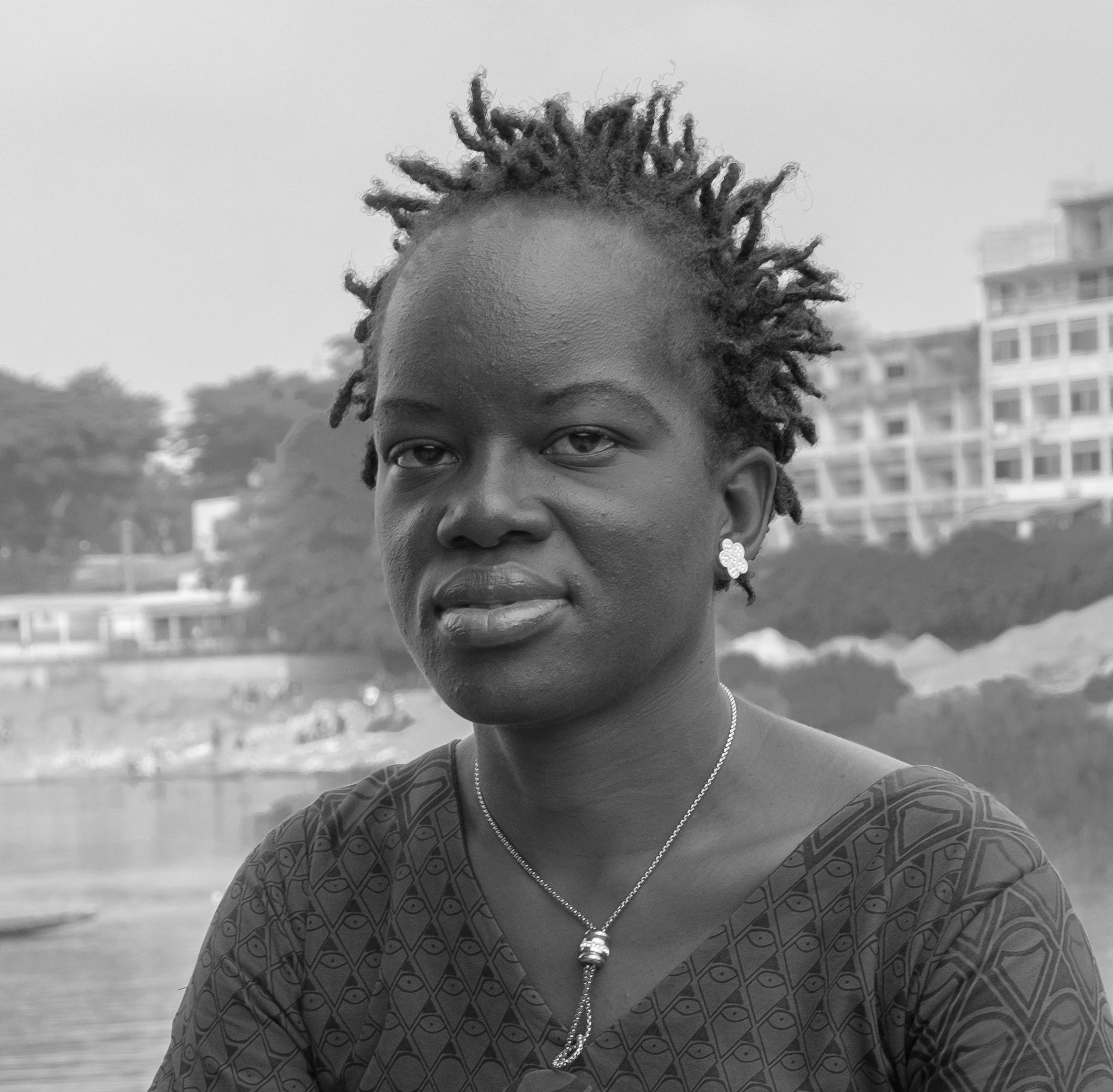 Pascale Appora-Gnekindy looks towards the camera. She wears a dark yellow African dress and yellowish necklace and sits on the riverbank.