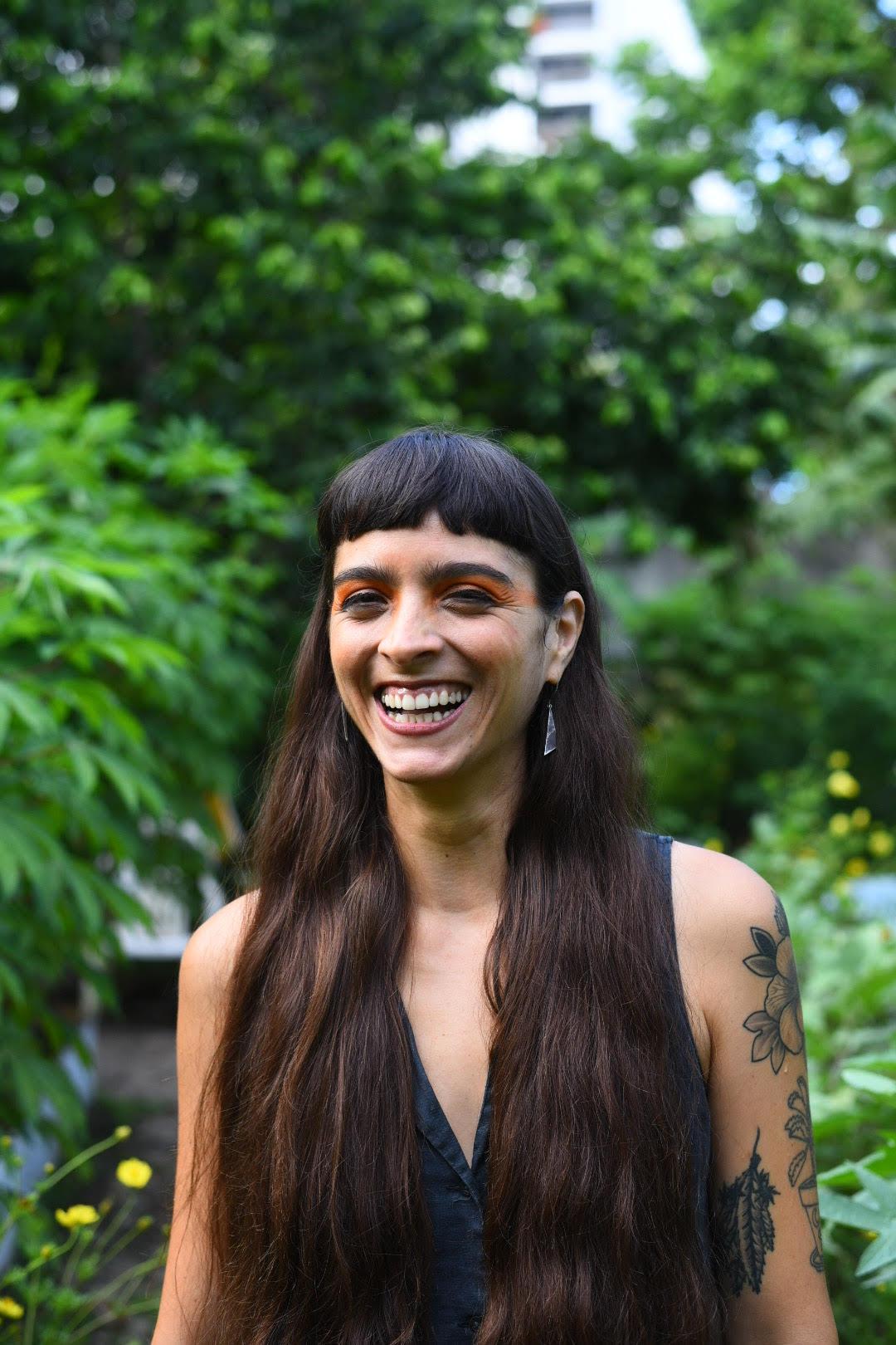 One of the producers for Matininó with long brown hair, bangs, orange eyeshadow and tattoos, Karla Claudia Betancourt, smiles for a photograph behind a backdrop trees.