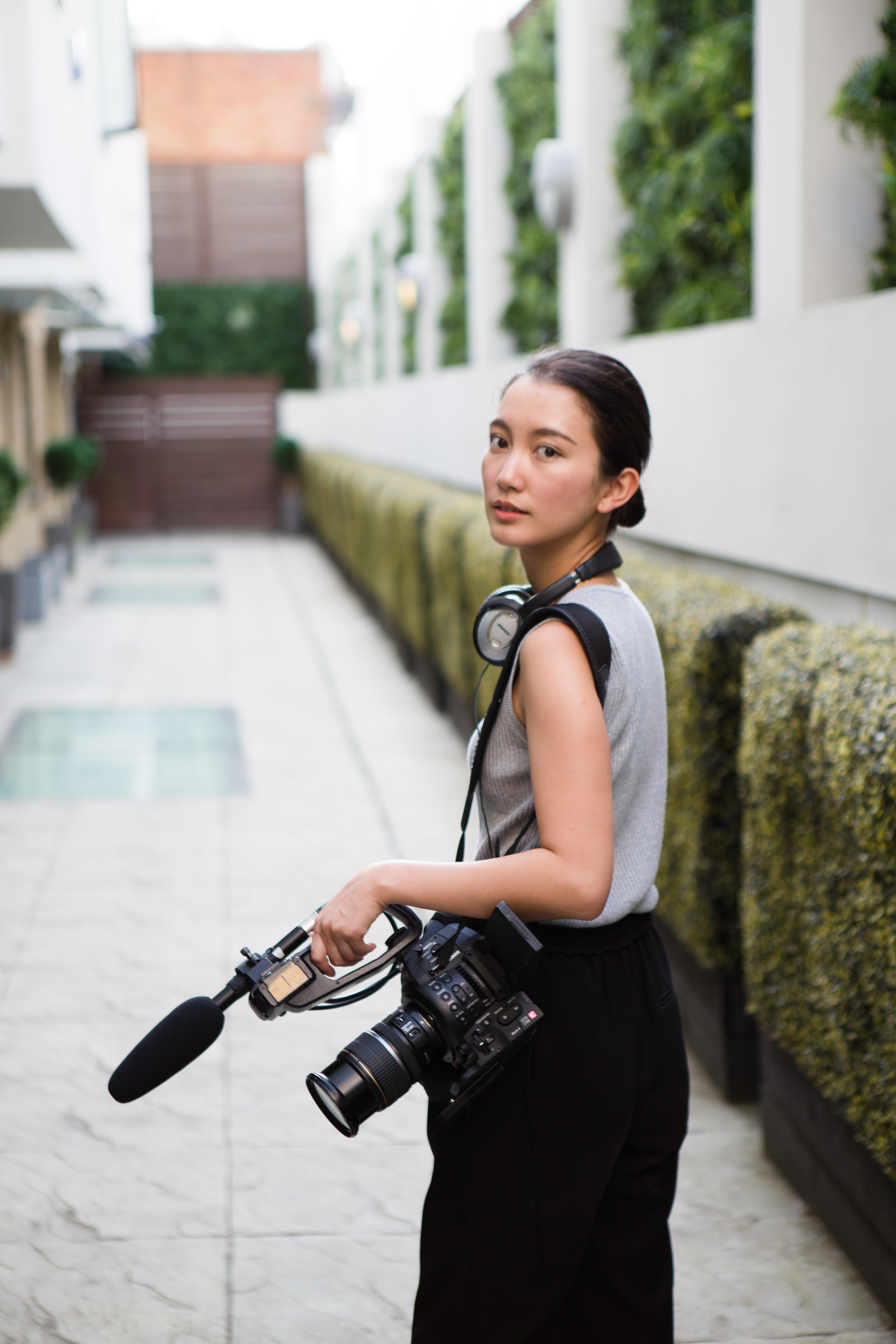 A woman, dark brown hair, standing outside with a film camera over her left shoulder, she wears headphones around her neck.