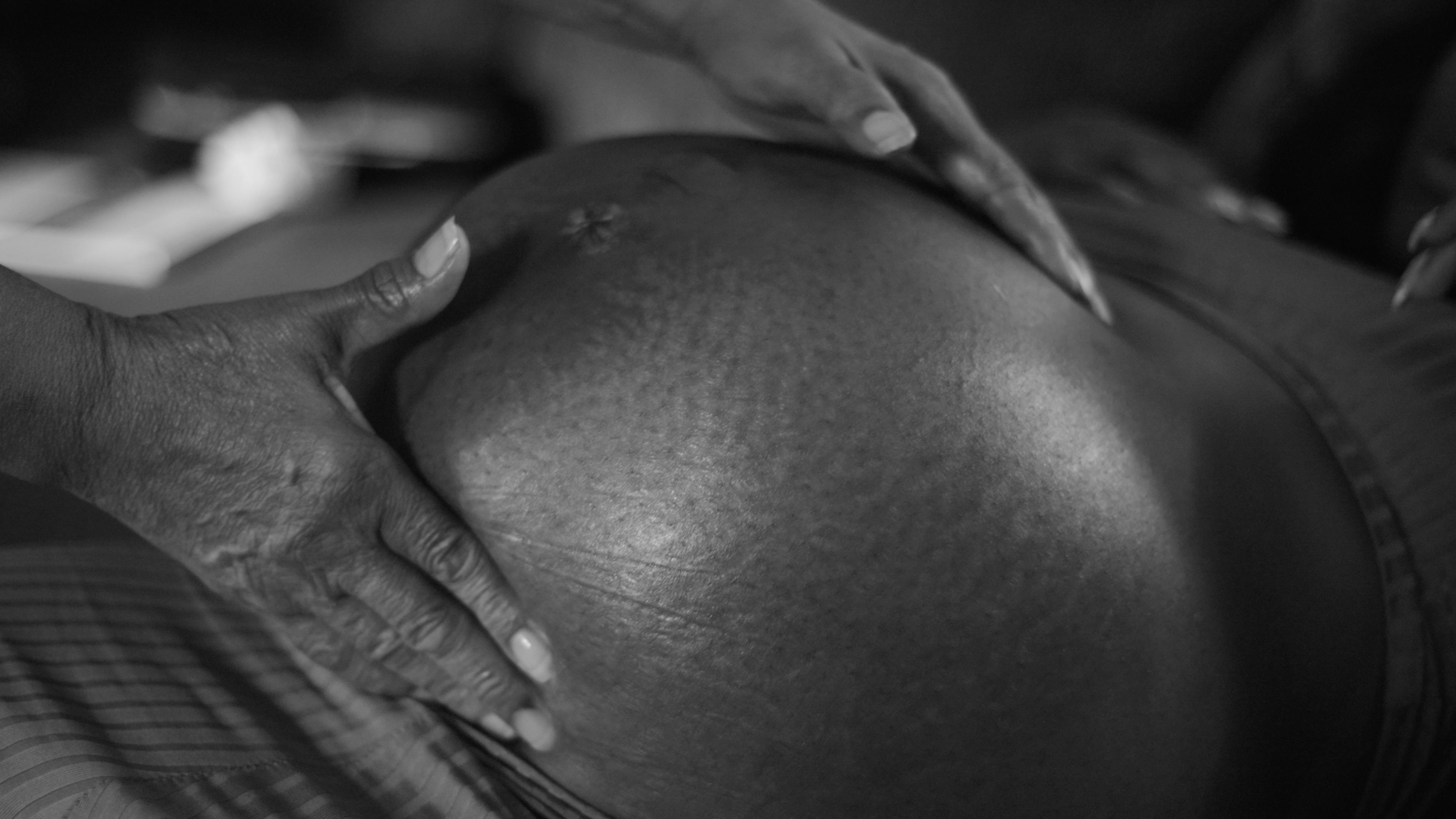 A close-up, black and white image showing a pregnant belly being gently held and examined by two pairs of hands.