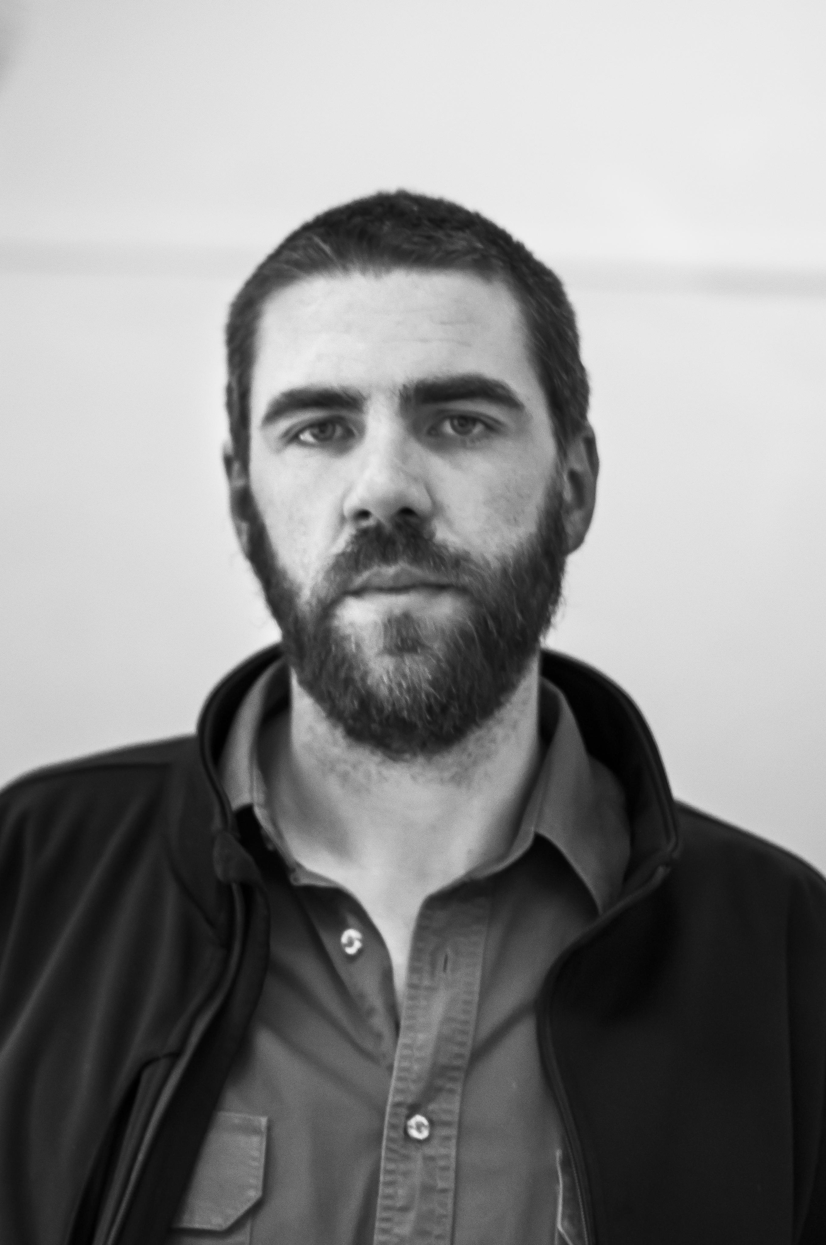 Headshot of a man with short hair, beard, a polo shirt and a jacket, looking at the camera