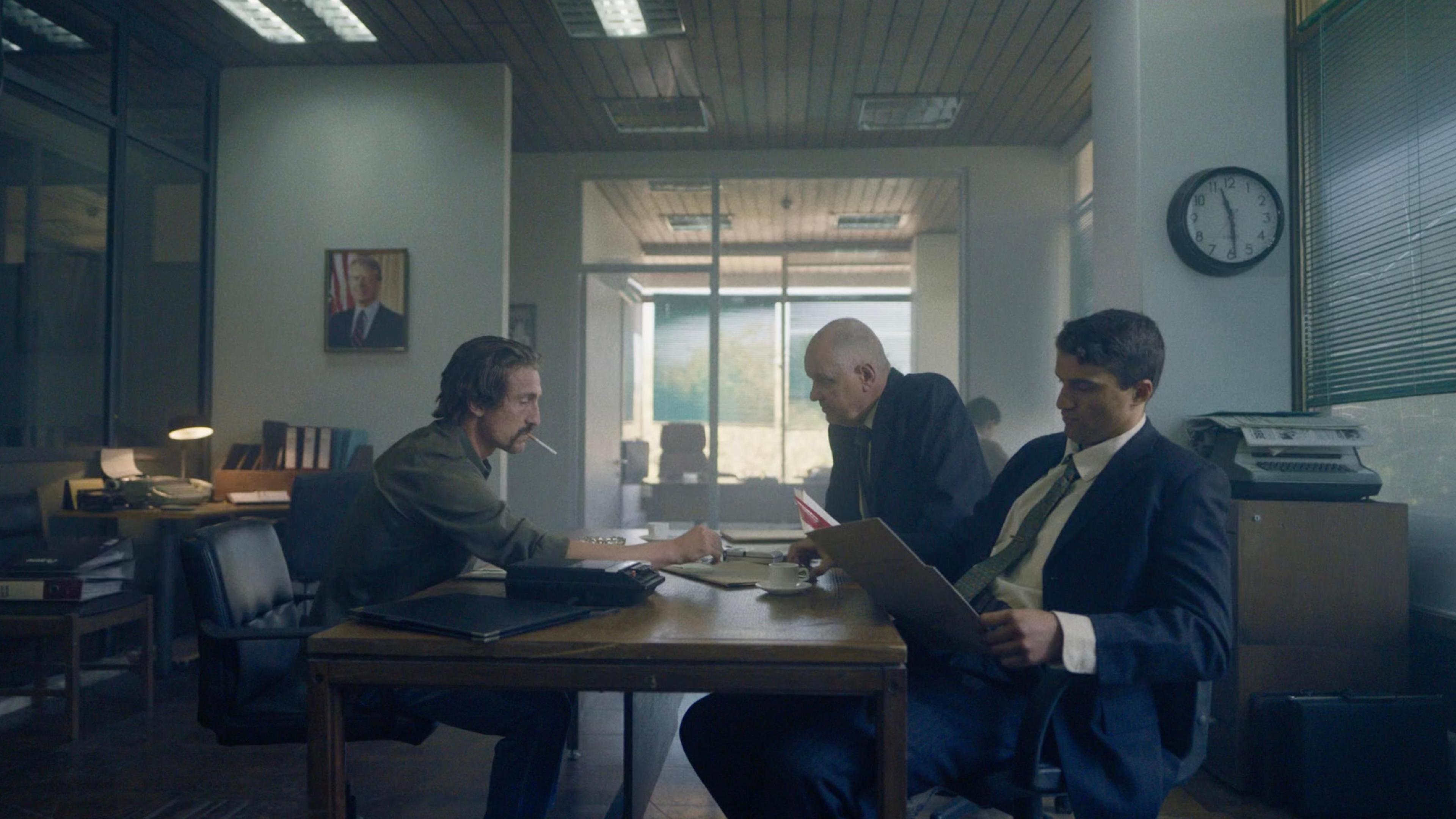 Three men sit at a table in an office building. One is smoking a cigarette, one is holding open a folder, and the other is leaning over the table.