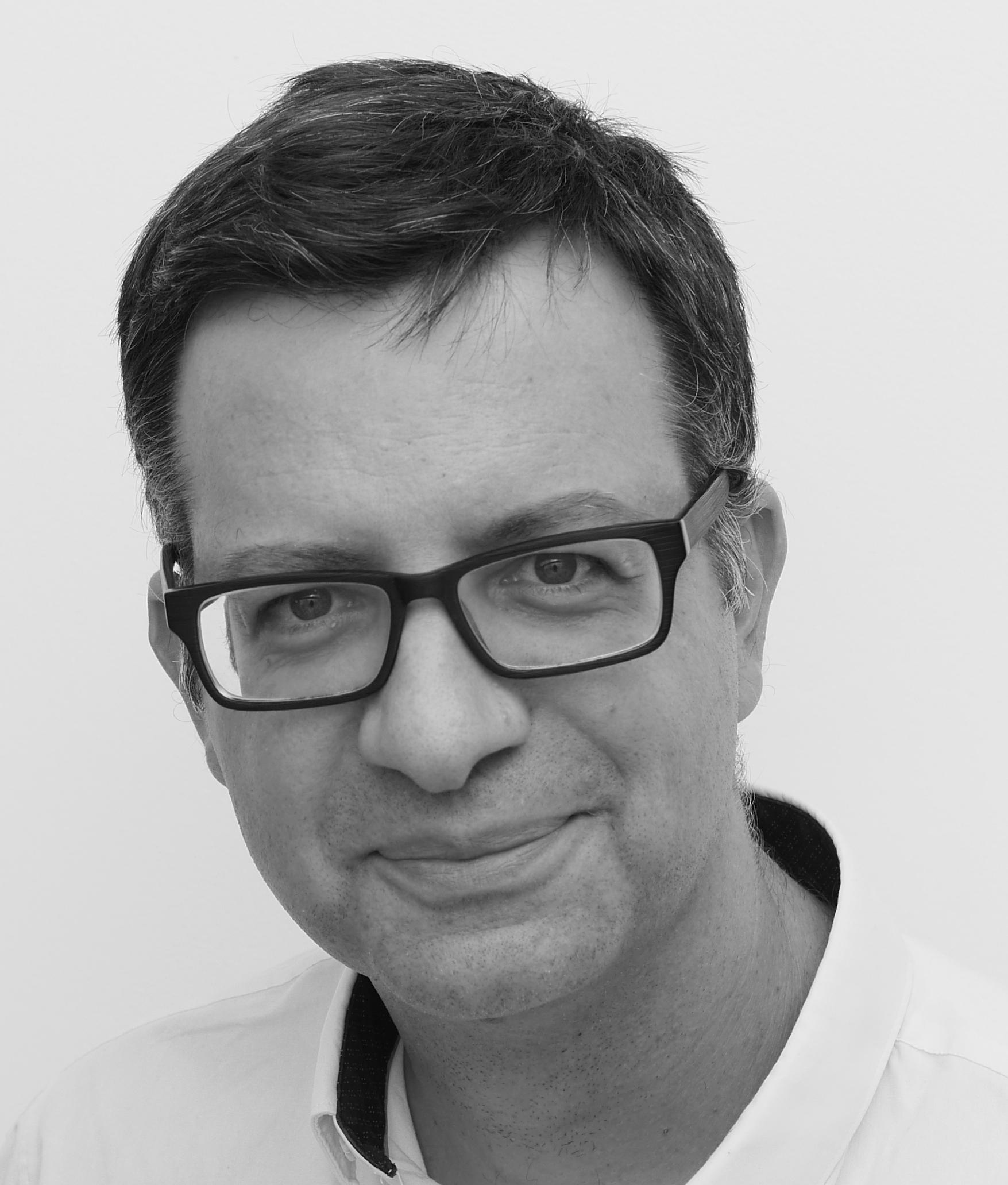 Headshot of a man wearing thick frames, short hair, looking directly at the camera and slightly smiling.