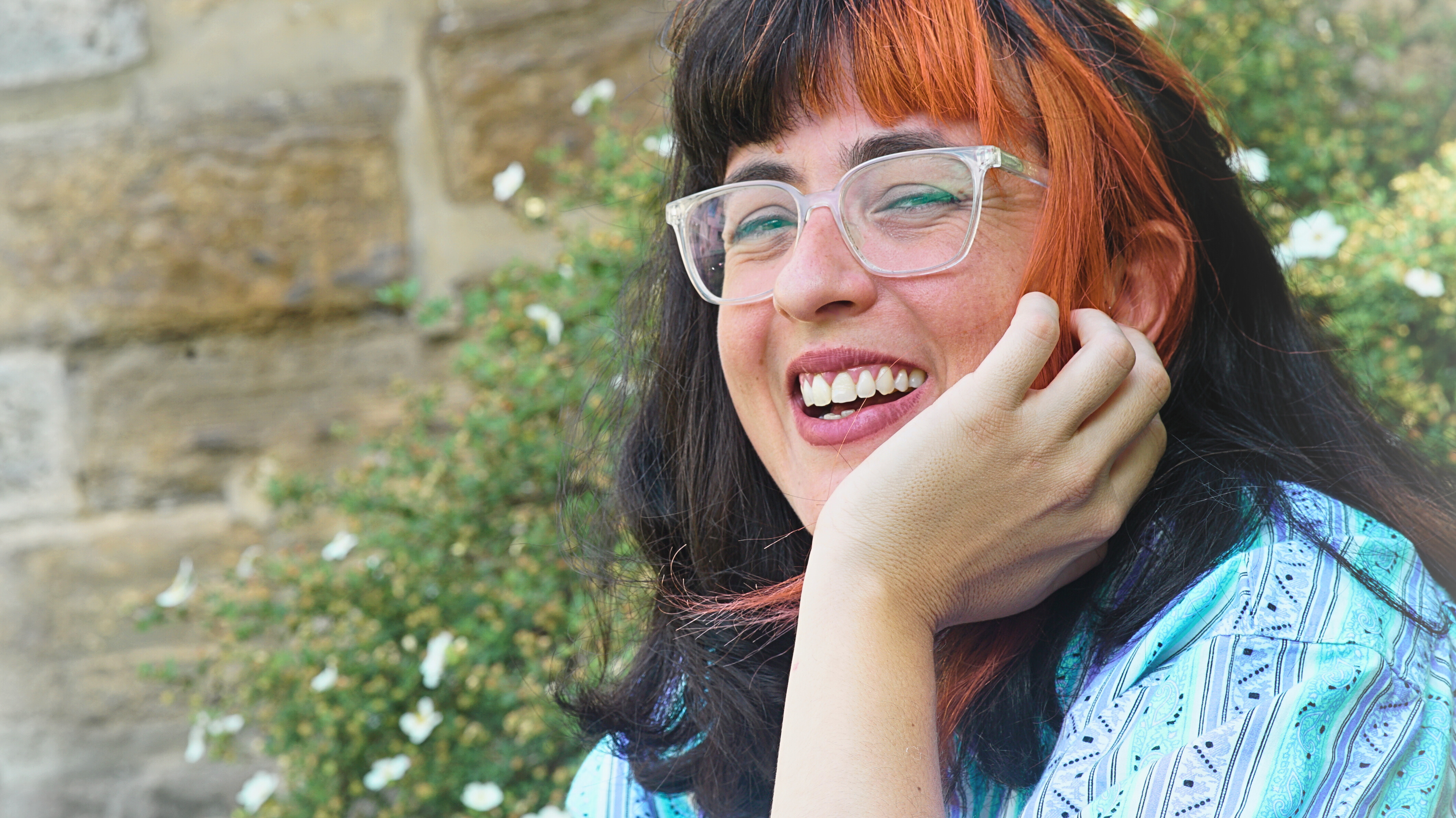 A woman with long dark hair and an orange streak smiles brightly while resting her head on her hand. She is wearing clear-framed glasses and a patterned turquoise shirt. The background features a stone wall and green shrubbery with small white flowers.