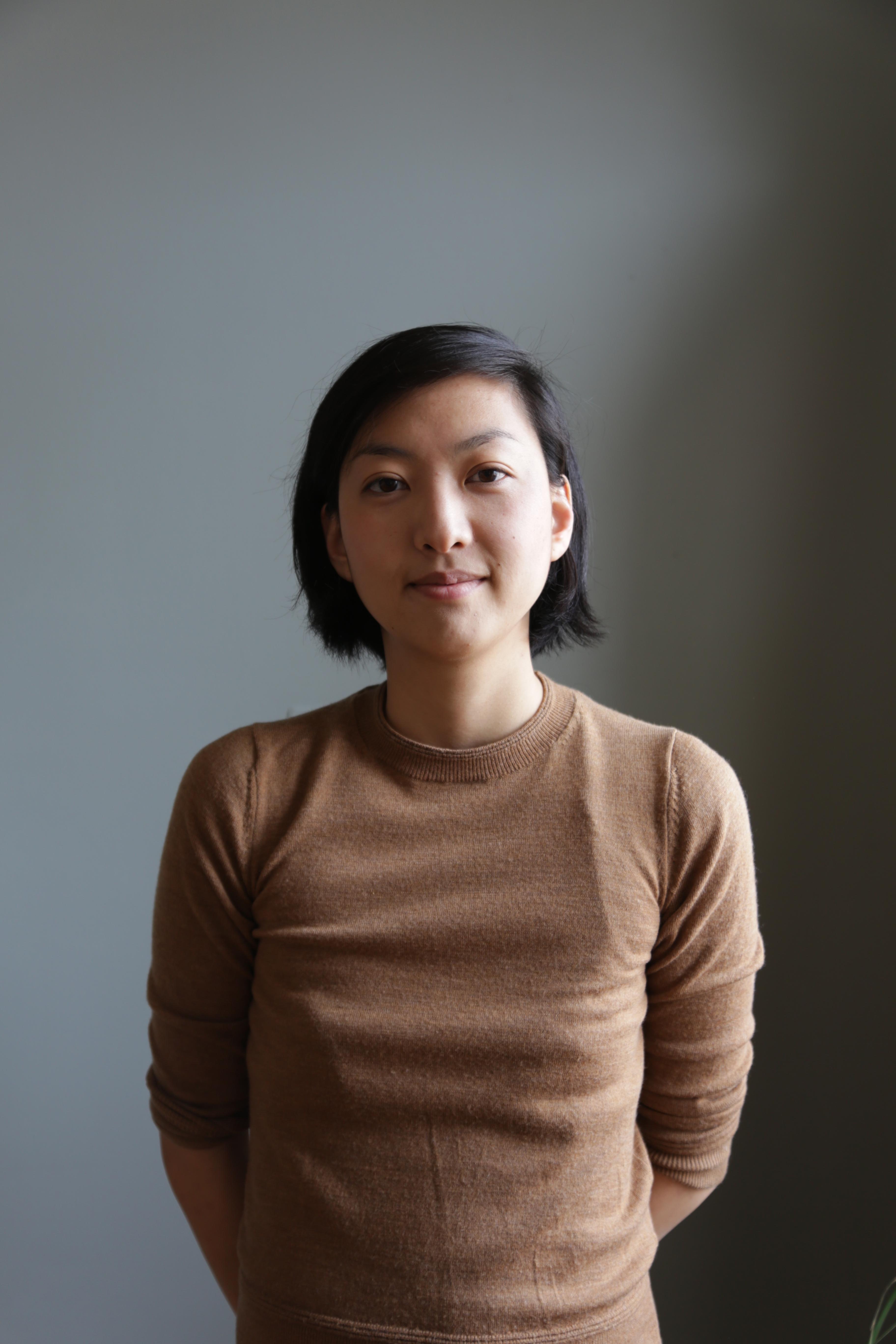A young, Taiwanese-American woman with chin-length black hair is wearing a brown sweater and standing in front of a gray wall.