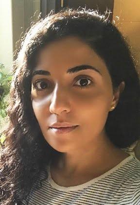 A woman with long, curly dark hair wearing a black and white striped shirt stands against an orange wall.