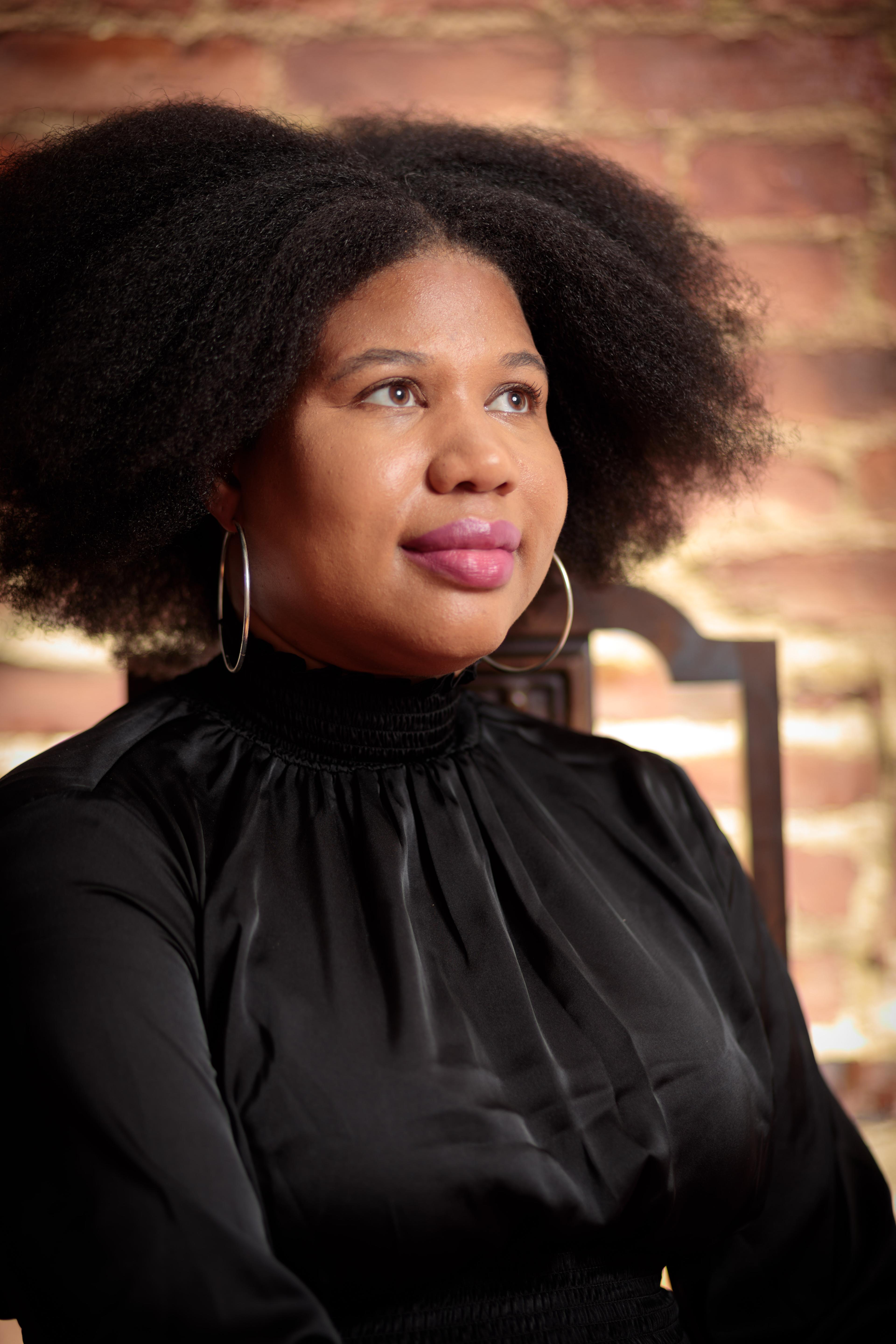 Princess Hairston looking off camera to her right. She has dark curly hair parted in the middle. She is wearing a black turtleneck blouse and large hoop earrings and is sitting in front of an out of focus brick wall.