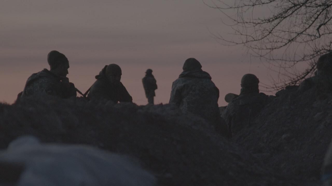 Still from My Dear Theo. We see soldiers sitting on the ground on the edge of a trench. We see only their backs as they are looking towards pink, evening sky. Only one of the soldiers is standing up, observing the horizon.