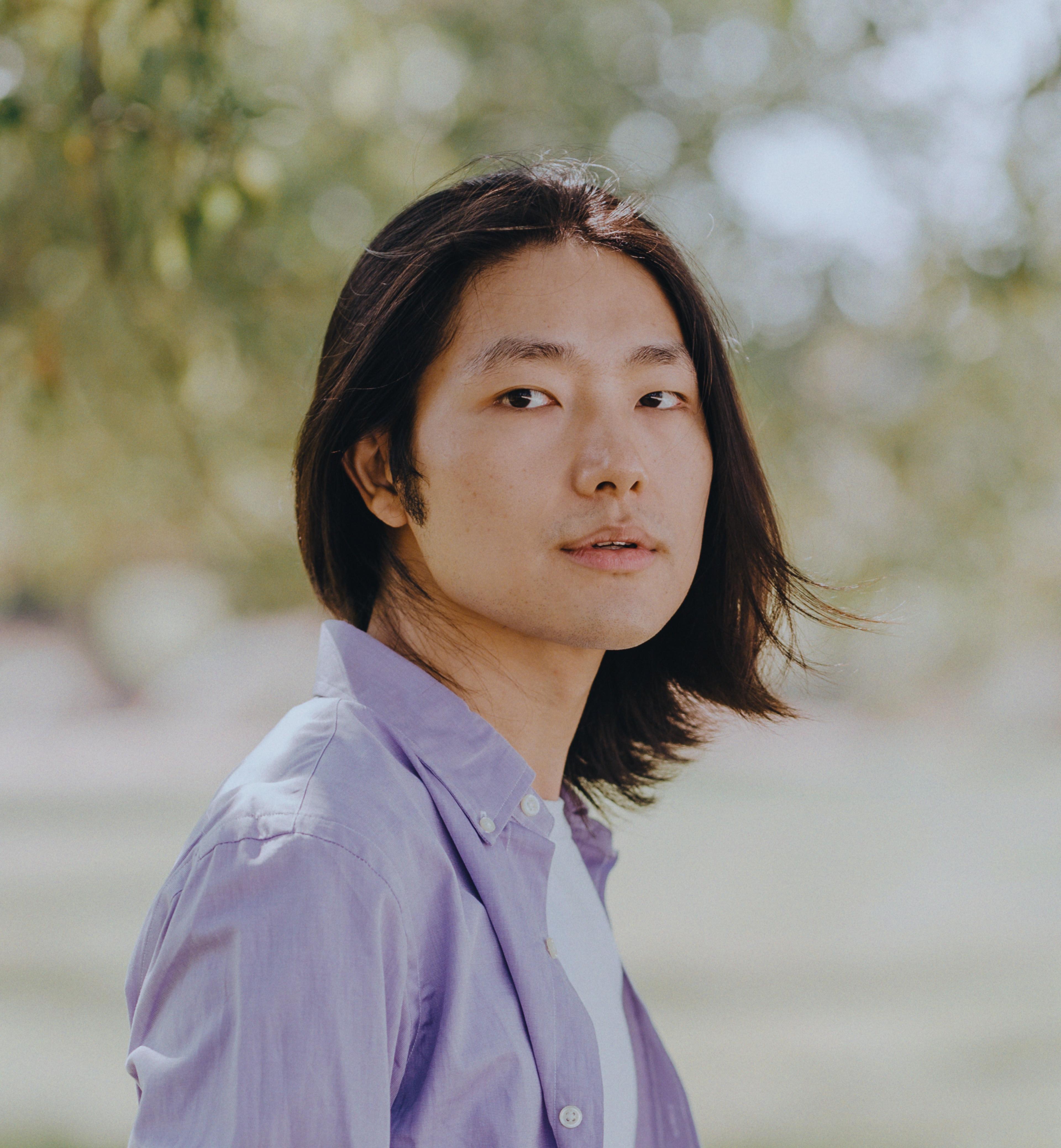 Headshot of a person with neck-length black hair, wearing a light purple shirt. They are facing the camera with a neutral expression, against an arboreal background.