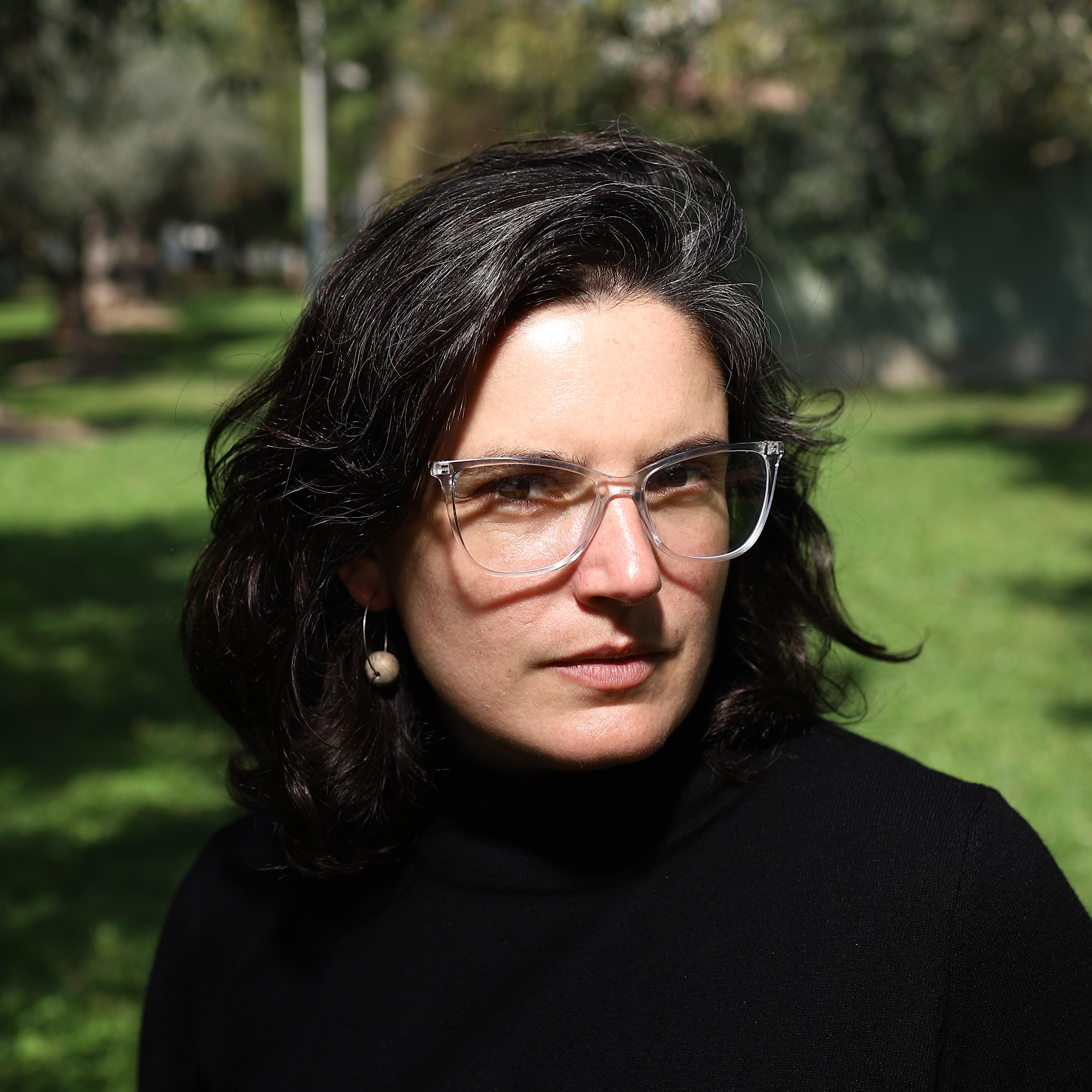 Portrait of filmmaker Agustina Pérez Rial outdoors, wearing a black turtleneck and clear-framed glasses, with a green park in the background.