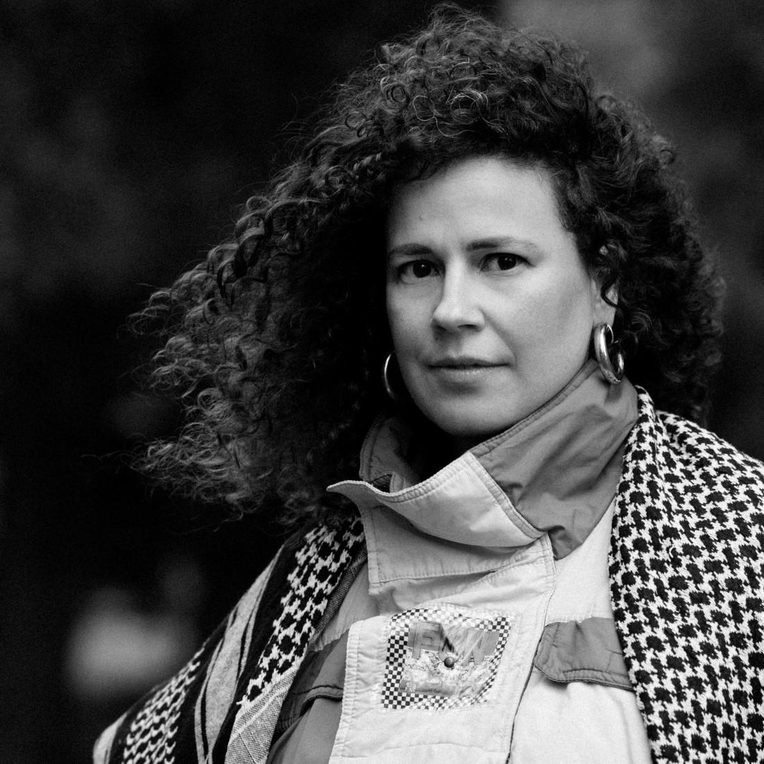 Black and white portrait of woman with curly hair wearing thick hoop earrings, and a coat with a turtle neck.