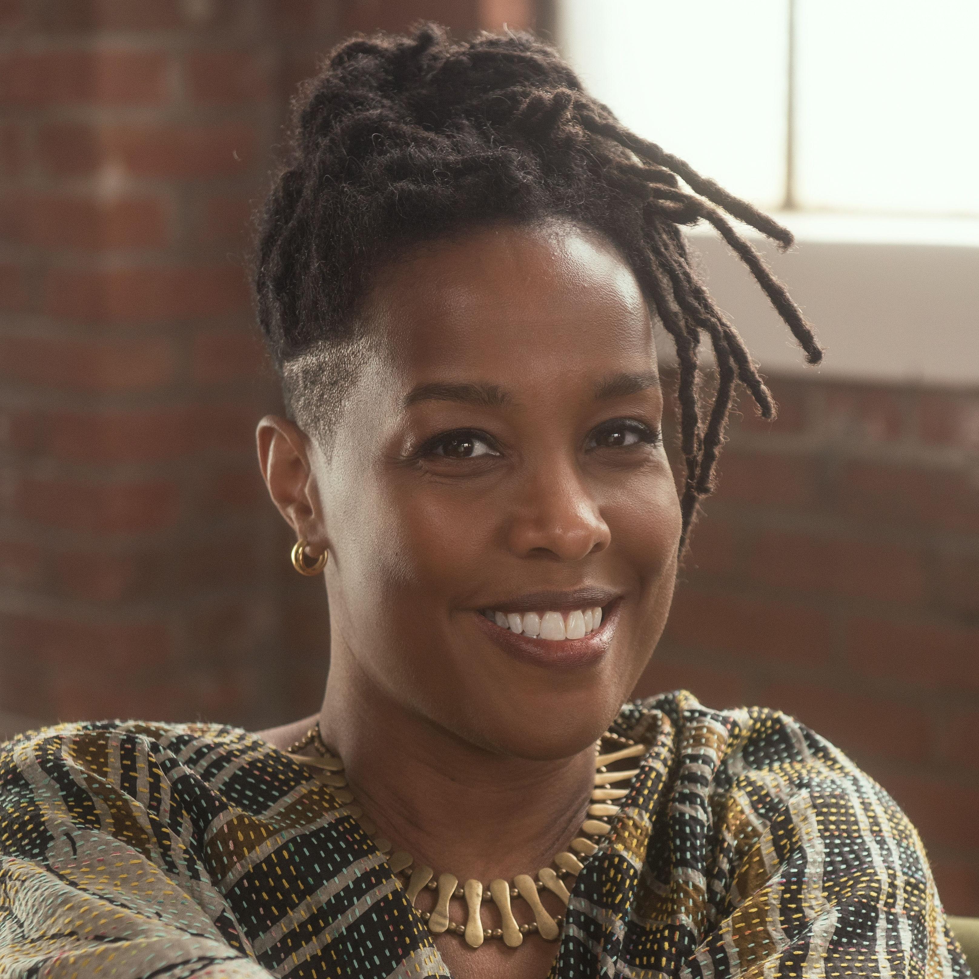 A woman sits on a green couch in front of a brick wall and smiles at the camera. She has her hair tied up and is wearing gold jewelry and a kantha top.