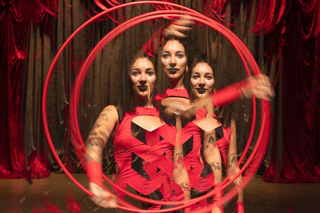 Midshot Image of woman wearing a red circus costume, black lipstick and playing with a red hoola hoop that frames her in the middle. The photo is stylistically edited to look kaleidoscopic.