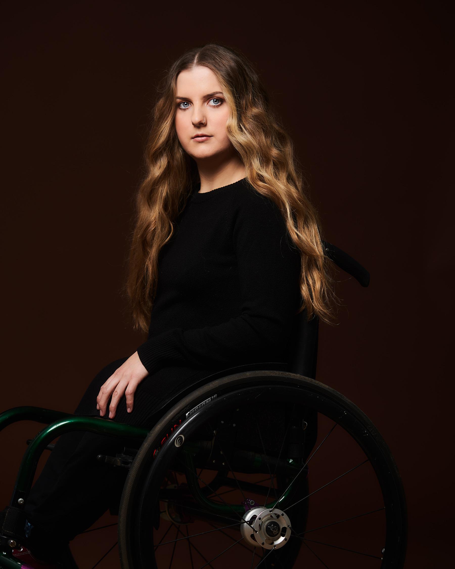 Ella Glendining looking straight ahead. She has light-colored, wavy, waist-length hair. She is wearing dark clothes and is sitting in a wheelchair.