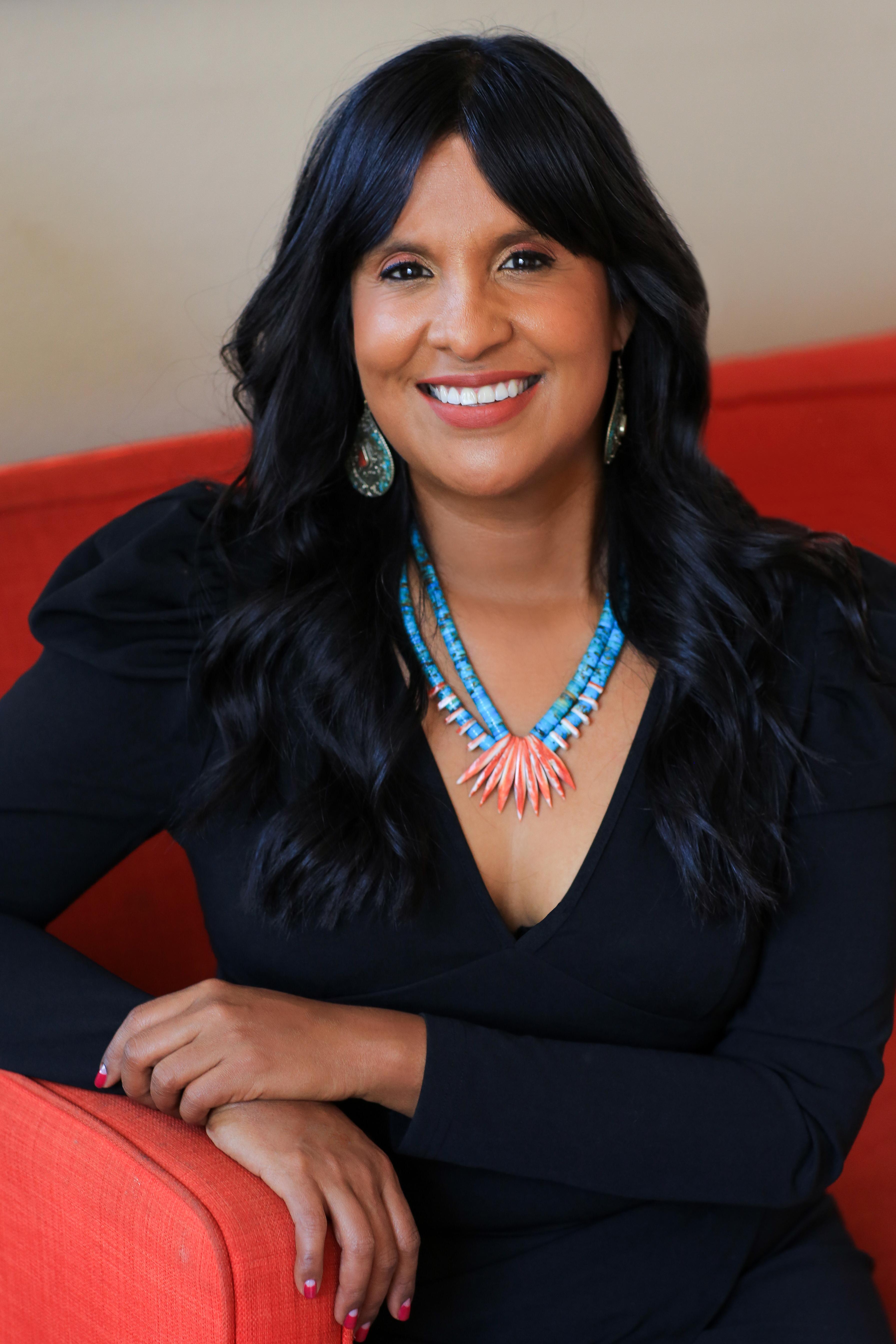A woman with long dark hair and decorative jewelry smiles into the camera.