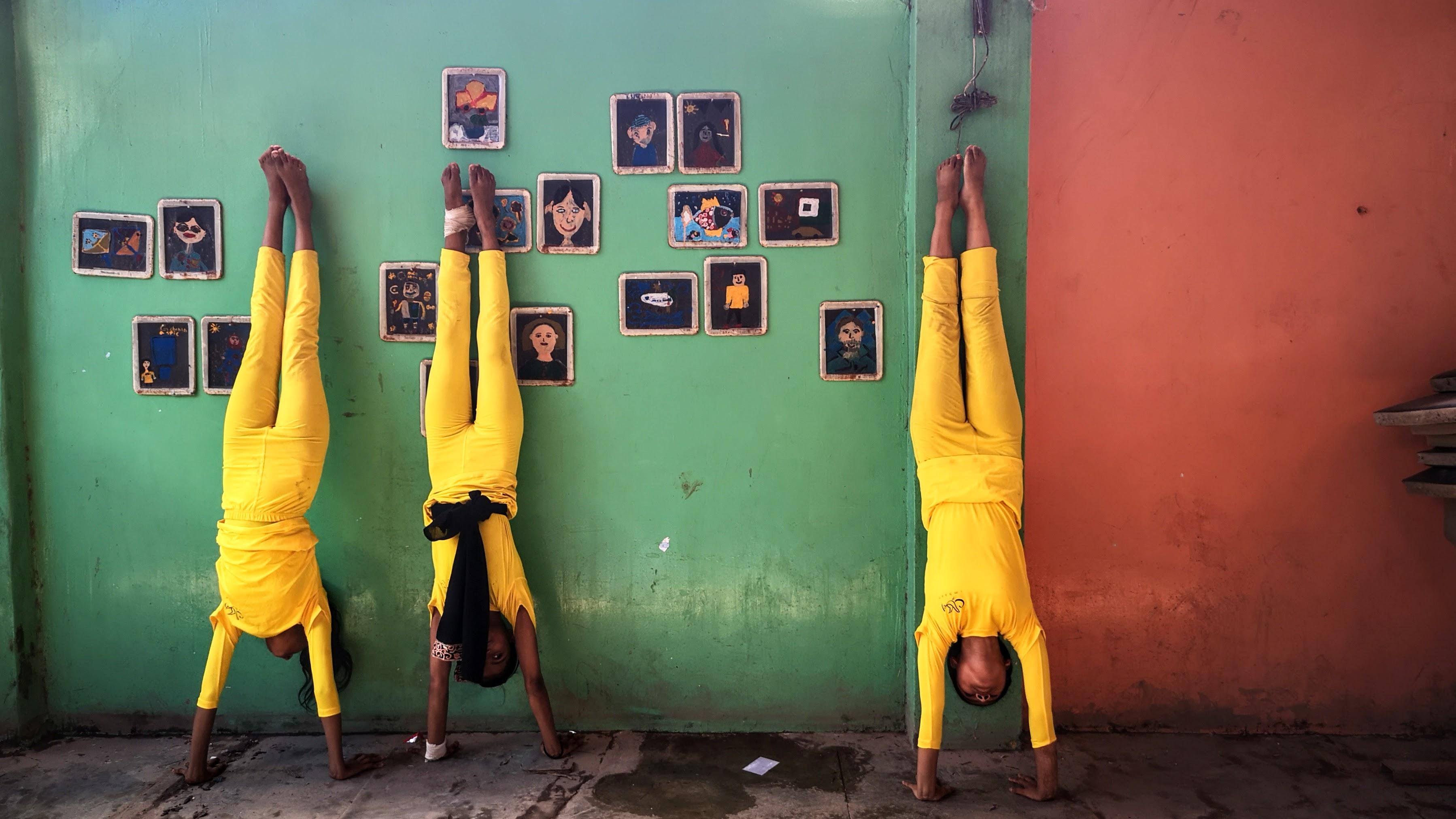 Three girls wearing matching yellow jumpsuits do headstands against a color-blocked green and red wall.