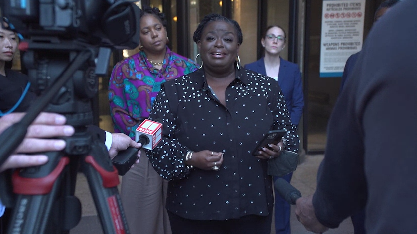 Monica Simpson, Executive Director of SisterSong, speaking at a press conference after the first day of the SisterSong v Georgia trial. She is directly facing the camera and is wearing hoop earrings and a black blouse with gold polka dots.