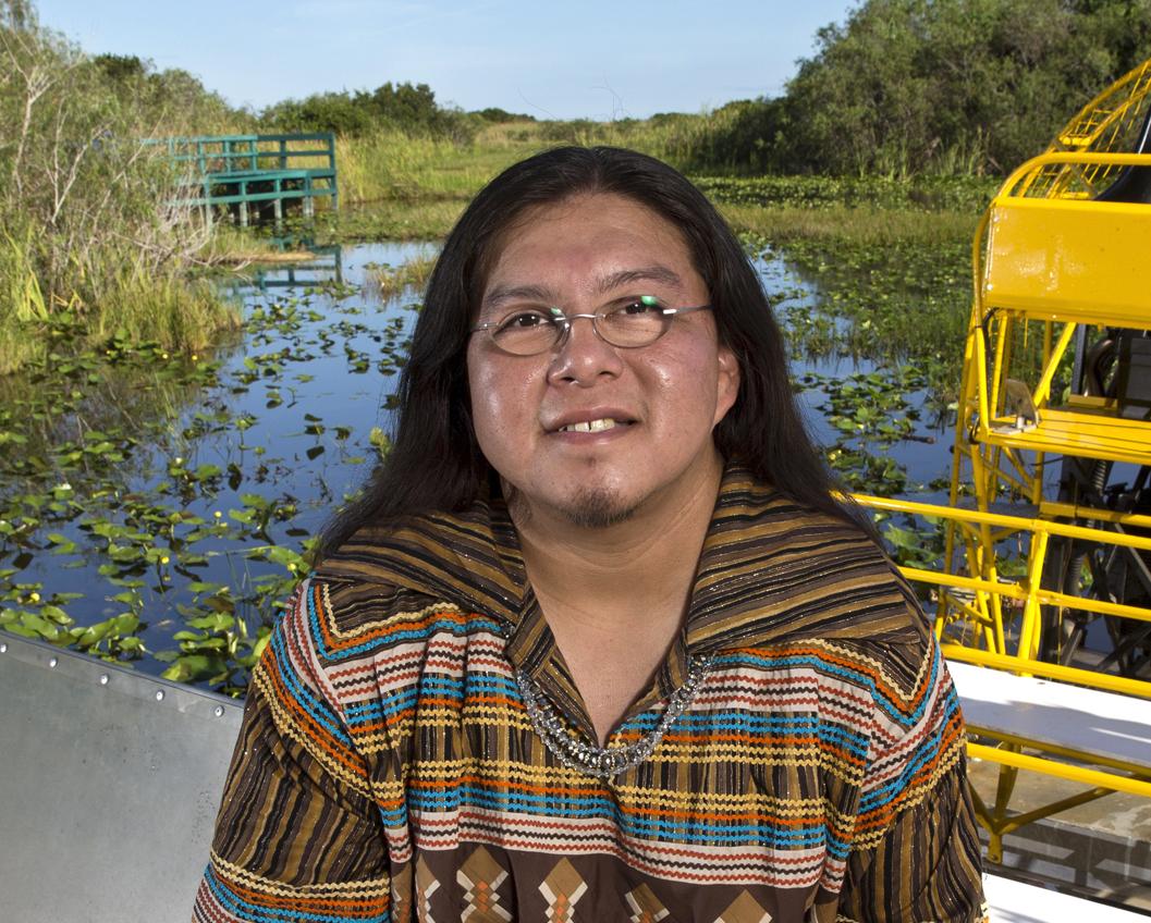Houston, a two spirit member of the Miccosukee Tribe, has long dark hair, glasses, and is wearing a Miccosukee patchwork top